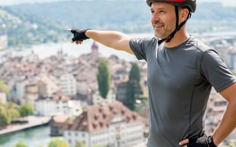 Lächelnder Radfahrer mit rotem Helm und grauem Shirt zeigt über malerische Schweizer Seestadt mit traditionellen Gebäuden und Bergen im Hintergrund.