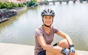 Lächelnde Radfahrerin mit Helm sitzt am Fluss mit historischer Brücke und Stadtpanorama im Hintergrund. Ausflugsboote auf dem Wasser.