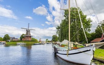 Weißes Segelboot am Kanal in Fryslan mit traditioneller Windmühle im Hintergrund unter blauem Himmel mit weißen Wolken.