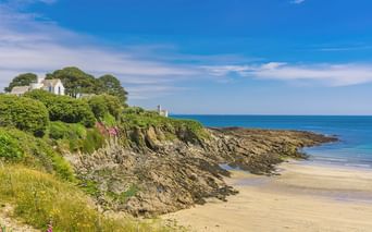 Sandstrand in Cornwall mit dunklen Felsenklippen und grüner Vegetation. Weiße Gebäude und Bäume auf der Klippe unter blauem Himmel.