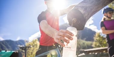 Radfahrer im roten Shirt füllt Wasserflasche an hölzernem Brunnen, mit Bergen und blauem Himmel im Hintergrund. Ein weiterer Radfahrer ist sichtbar.