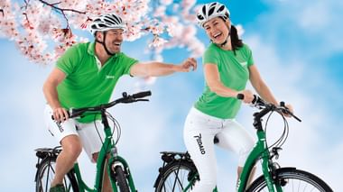Zwei Radfahrer in grünen Pedalo-Shirts und weißen Helmen fahren grüne Fahrräder unter rosa Kirschblüten vor blauem Himmel.