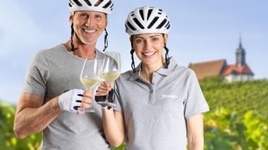 Zwei lächelnde Radfahrer mit Helmen stoßen mit Weißweingläsern in einem Weinberg an. Ein Kirchturm ist im Hintergrund sichtbar.