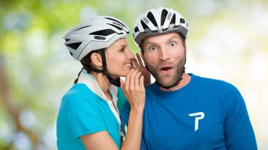 Frau in türkisfarbenem Shirt und weißem Helm flüstert überraschtem Mann in blauem Shirt zu. Beide tragen weiße Fahrradhelme vor unscharfem Hintergrund.
