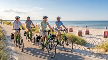 Vier Radfahrer mit Helmen und Packtaschen auf grünen Rädern auf einem Küstenweg. Sandstrand mit Strandkörben und ruhiger See im Hintergrund.