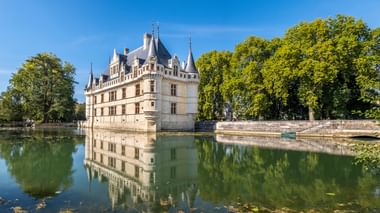 Weißes Renaissance-Schloss mit spitzen Türmchen spiegelt sich im umgebenden Wassergraben, eingerahmt von grünen Bäumen unter blauem Himmel.