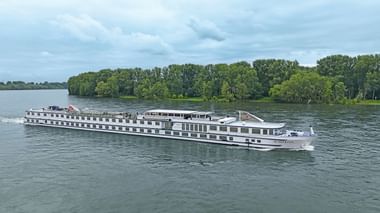 Weißes Flusskreuzfahrtschiff Rigoletto fährt auf ruhigem Wasser mit grünen bewaldeten Ufern im Hintergrund unter bewölktem Himmel.