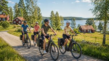Four cyclists riding on a gravel road past red wooden houses with a lake and forest in the background under a blue sky in Scandinavia.