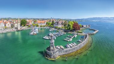 Aerial view of Lindau harbor on Lake Constance with lighthouse, marina, historic buildings with red roofs, and turquoise water under blue sky.