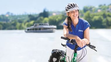 Lächelnde Frau mit Helm auf Fahrrad am Fluss. Ausflugsschiff und grüne Landschaft im Hintergrund unter blauem Himmel sichtbar.
