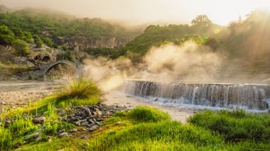 Historische Steinbogenbrücke und Wasserfall in nebelverhangener albanischer Landschaft mit grünen Hügeln, fließendem Fluss und Morgenlicht.