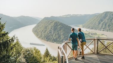 Zwei Radfahrer auf einer Aussichtsplattform mit Blick auf die Donau, die sich durch bewaldete Hügel schlängelt. Ein Frachtschiff ist auf dem Fluss sichtbar.