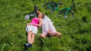 Zwei Radfahrer liegen auf grünem Gras mit einem türkisfarbenen Fahrrad neben ihnen. Sie tragen Radkleidung und Helme sind in der Nähe sichtbar.