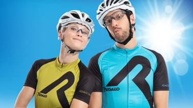 Zwei Radfahrer mit weißen Helmen und Brillen posieren vor blauem Himmel. Einer trägt ein gelbes, der andere ein türkisfarbenes Trikot.
