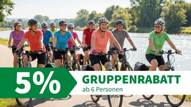 Gruppe von Radfahrern mit Helmen und bunten Shirts auf einem asphaltierten Weg neben einem Fluss mit Bäumen im Hintergrund.