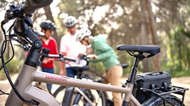 Die Freude am gemeinsamen Radeln steht bei diesen Reisen im Vordergrund, da sind sich alle einig (Bild: winora.de, pd-f) Nahaufnahme eines weißen E-Bikes mit Winora-Marke im Vordergrund, mit unscharfer Radfahrergruppe mit Helmen im Hintergrund zwischen Bäumen.