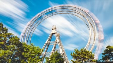 Langzeitbelichtung des Wiener Prater Riesenrads in Bewegung, mit kreisförmiger Unschärfe vor blauem Himmel, umgeben von grünen Bäumen.