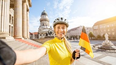 Lächelnde Radfahrerin in gelbem Trikot und Helm mit deutscher Flagge am Gendarmenmarkt in Berlin mit Französischem Dom im Hintergrund.
