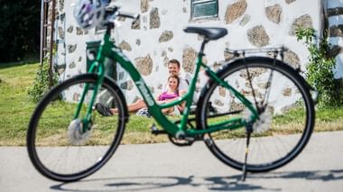 Grünes Fahrrad im Vordergrund mit zwei Radfahrern, die auf der Wiese vor einem weißen Steingebäude am Meridiem Trail rasten.