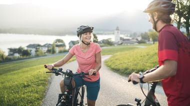 Zwei Radfahrer mit Helmen auf einem asphaltierten Weg in Oberösterreich. Grüne Wiesen und ein Seeufer-Dorf mit Kirchturm im Hintergrund.