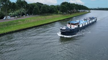Aerial view of hotel barge Magnifique 4 with blue and white hull cruising on a canal. Tree-lined banks and parked cars visible on the left shore.