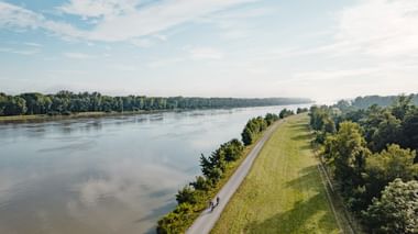 Luftaufnahme von zwei Radfahrern auf einem asphaltierten Weg entlang eines breiten Flusses. Grüner Damm trennt den Weg vom Wasser, Wälder auf beiden Seiten.