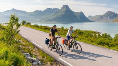 Two cyclists with panniers riding on a coastal road in Norway with dramatic mountain peaks and fjord in the background under blue sky.