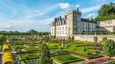 Weißes Steinschloss mit Turm und formalen Gärten mit geometrischen Hecken, bunten Blumenbeeten und gepflegten Rasenflächen unter blauem Himmel.