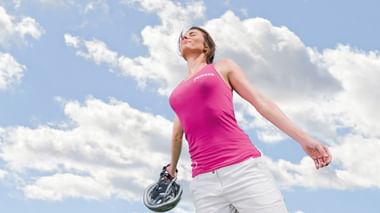 Frau in pinkem Radtrikot und weißer Shorts hält einen Helm, steht vor blauem Himmel mit weißen Wolken, Augen geschlossen, genießt den Moment.