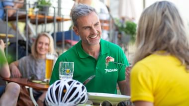 Mann im grünen Pedalo-Poloshirt mit Tablet spricht mit Radfahrerin im gelben Shirt im Straßencafé. Fahrradhelm auf dem Tisch sichtbar.