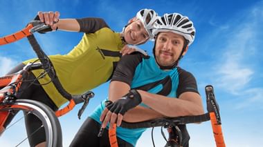 Zwei lächelnde Radfahrer mit Helmen und Radkleidung posieren mit ihren Rädern vor blauem Himmel. Einer trägt ein gelbes, der andere ein türkises Trikot.