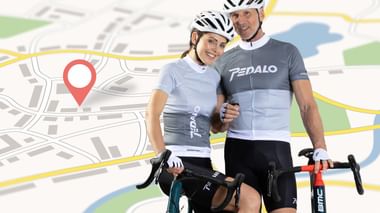 Two smiling cyclists in helmets and cycling jerseys holding road bikes, standing in front of a stylized map with a red location pin.