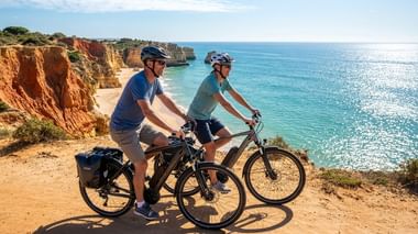 Two cyclists with e-bikes on sandy cliff path overlooking turquoise ocean and golden rock formations along the Algarve coast.
