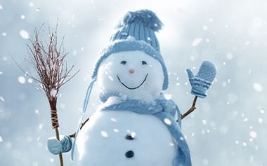 Fröhlicher Schneemann mit blauer Strickmütze und Schal, hält Reisigbesen und winkt mit blauem Fäustling. Schnee fällt vor grauem Winterhimmel.