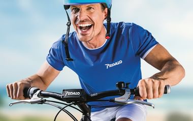 Mann mit blauem Helm und passendem Shirt lacht beim Radfahren im Freien. Das Pedalo-Logo ist auf Shirt und Fahrrad sichtbar.