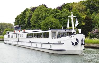 Weißes Flusskreuzfahrtschiff namens Fluvius mit großen Panoramafenstern fährt auf ruhigem Wasser, umgeben von üppigen grünen Bäumen am Flussufer.