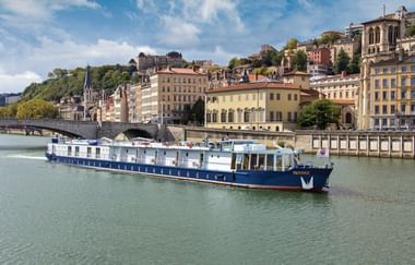 Blau-weißes Flusskreuzfahrtschiff auf ruhigem Wasser mit historischen französischen Stadtgebäuden und Brücke im Hintergrund unter bewölktem Himmel.