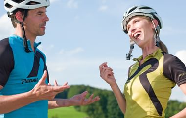 Mann in blauem Radtrikot und Frau in gelbem Trikot mit Helmen sprechen im Freien vor blauem Himmel und grüner Landschaft.