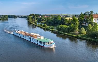 Weißes Flusskreuzfahrtschiff Florentina fährt auf ruhigem Fluss, umgeben von grünen Bäumen und Landschaft mit Häusern am Ufer.