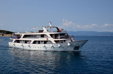 Weißes mehrstöckiges Passagierschiff Andela Lora auf ruhigem blauen Wasser mit bewaldeter Küste und Bergen im Hintergrund.