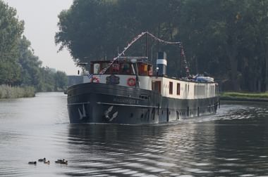 Hotel barge Magnifique II cruising on a tree-lined canal. The black-hulled vessel has white cabins and decorative lights. Ducks swim nearby.
