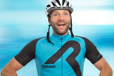 Lächelnder Radfahrer mit weißem Helm und blau-schwarzem Radtrikot mit großem Fragezeichen-Design vor türkis gestreiftem Hintergrund.
