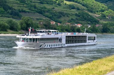 Weißes Flusskreuzfahrtschiff Lisabelle fährt auf einem Fluss mit grünem Hügeldorf im Hintergrund. Das moderne Schiff hat mehrere Decks.