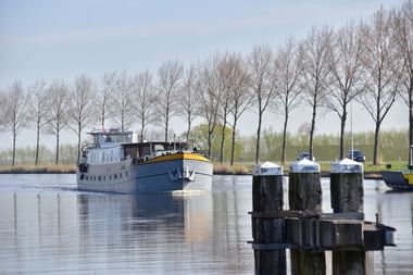 Grau-gelbes Tretboot Lena Maria auf ruhigem Wasser mit hölzernen Pollern im Vordergrund und kahlen Bäumen am Ufer.