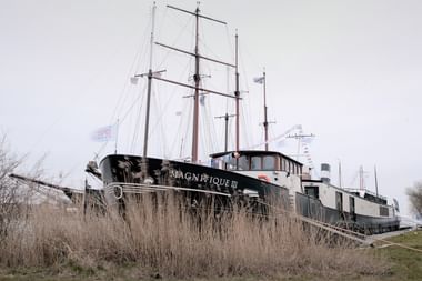 The Magnifique III, a dark-hulled vessel with three masts and white superstructure, moored behind dry reeds along a waterway under overcast sky.