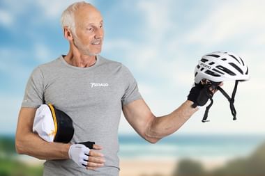 Lächelnder Mann im grauen Pedalo-Shirt hält zwei Fahrradhelme und trägt weiße Radhandschuhe vor unscharfem Küstenhintergrund.