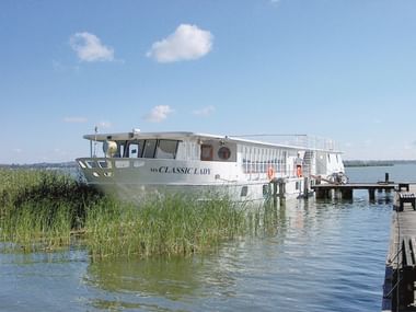 Weißes Passagierschiff MS Classic Lady an einem Holzsteg auf einem ruhigen See. Schilf wächst im flachen Wasser, blauer Himmel mit Wolken darüber.