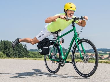 Begeisterter Radfahrer mit gelbem Helm und grünem Shirt fährt ein grünes Tourenrad auf einer Straße, ein Bein spielerisch ausgestreckt.