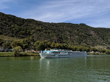 White and blue river cruise ship sailing on calm green water in Switzerland, with forested hills and small village in the background under blue sky.