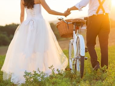 Bride in white wedding dress and groom in suspenders holding hands while walking with a white bicycle with basket in a sunny meadow.